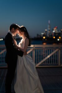 Couple embrace at blue hour with the Manhattan skyline glowing behind the Liberty Warehouse pier