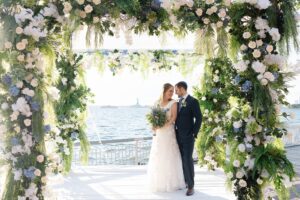 Bride and groom beneath their floral chuppah with the Statue of Liberty visible across the harbor