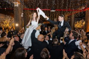 Bride and groom lifted in chairs holding a handkerchief during the hora at their Liberty Warehouse Jewish wedding